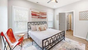 A bedroom with a bed, a ceiling fan, a window with blinds, a red chair, and a door leading to another room. - The Lair at Georgia Tech and Mercedes-Benz Stadium (Atlanta)