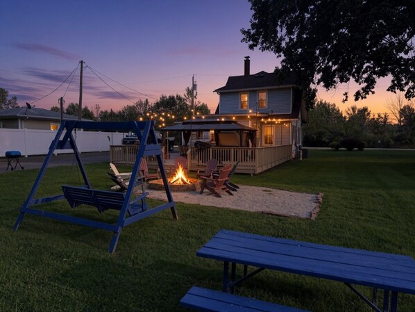 A backyard with a swing set, a fire pit, and a deck with string lights.