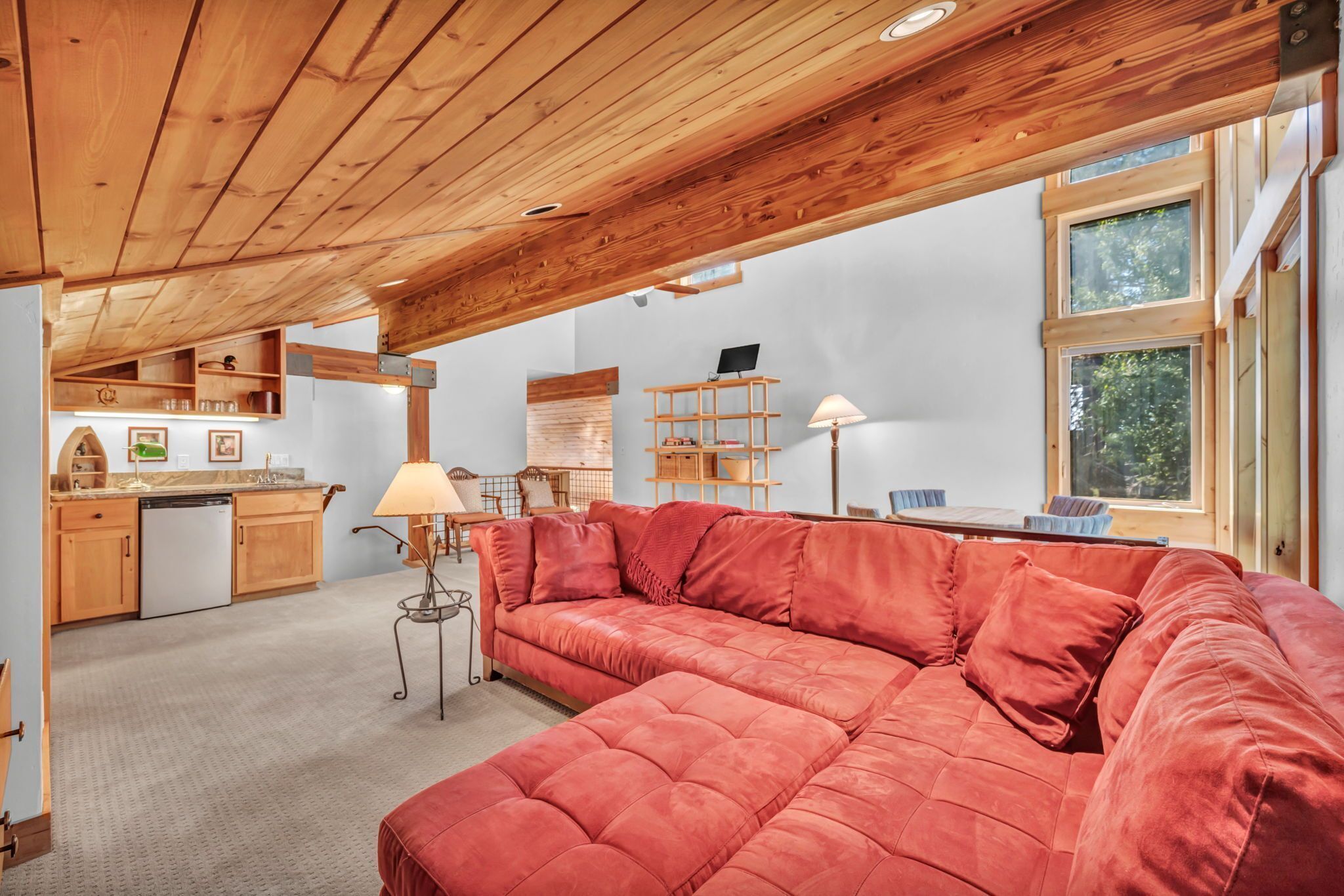 A cozy living area with a red sofa, wooden ceiling beams, a kitchen with wooden cabinets, and a window with a view of greenery.