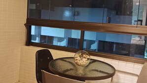 Een eetgedeelte met een ronde glazen tafel en twee stoelen, een groot raam met uitzicht op gebouwen, en witte betegelde wanden.