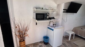 Een compacte keuken met een magnetron, een kleine koelkast en een spoelbak. Boven de magnetron is een plank met servies en glazen. Naast de koelkast staat een klein krukje, en op de vloer staat een potplant.