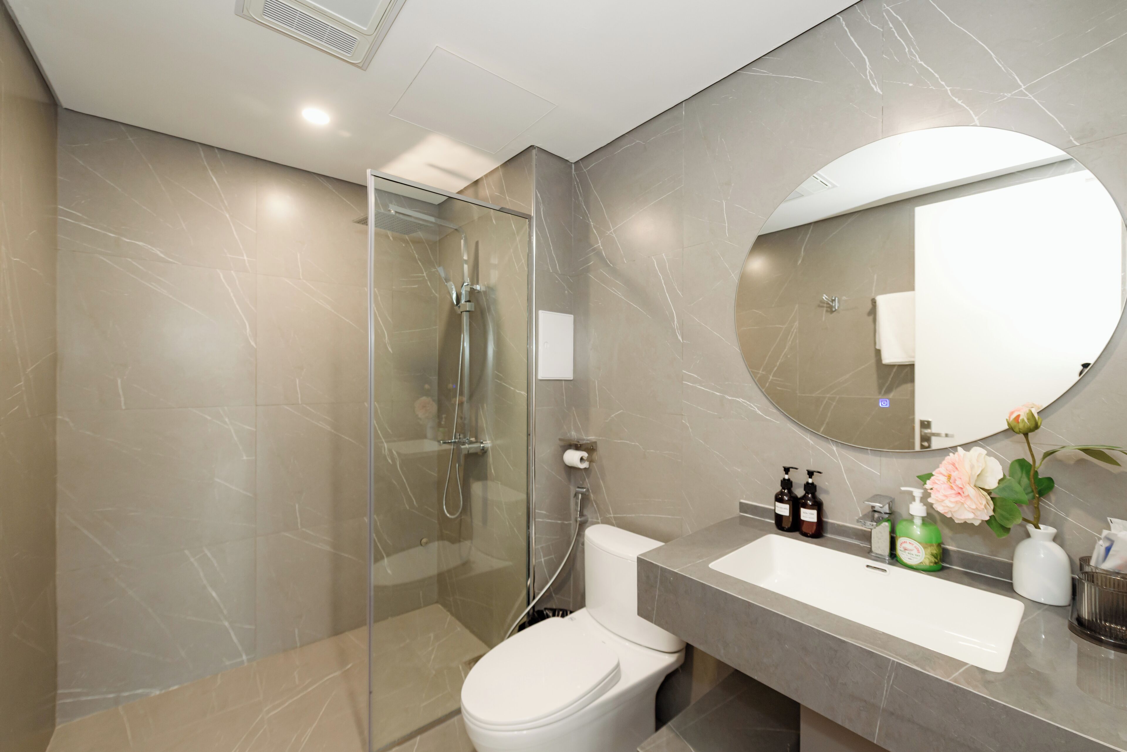 Een moderne badkamer met een douchecabine van glas, een wit toilet, een wand betegeld met marmer, een grote spiegel en een wastafel met toiletartikelen.