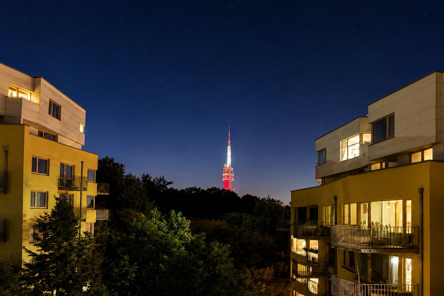 A nighttime view of a building with a prominent tower in the background, surrounded by trees and other buildings.
