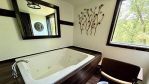 A modern bathroom with a large bathtub, a wooden vanity, a mirror, and a decorative wall piece. - Happy Hideaway in our charming cabin in historic Eureka Springs with jacuzzi tub (Eureka Springs)