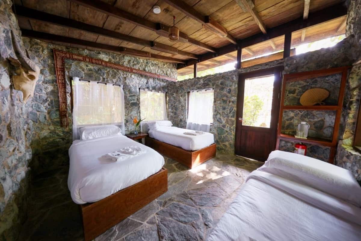 A rustic room with stone walls and wooden beams, featuring two beds with white linens and a stone floor.
