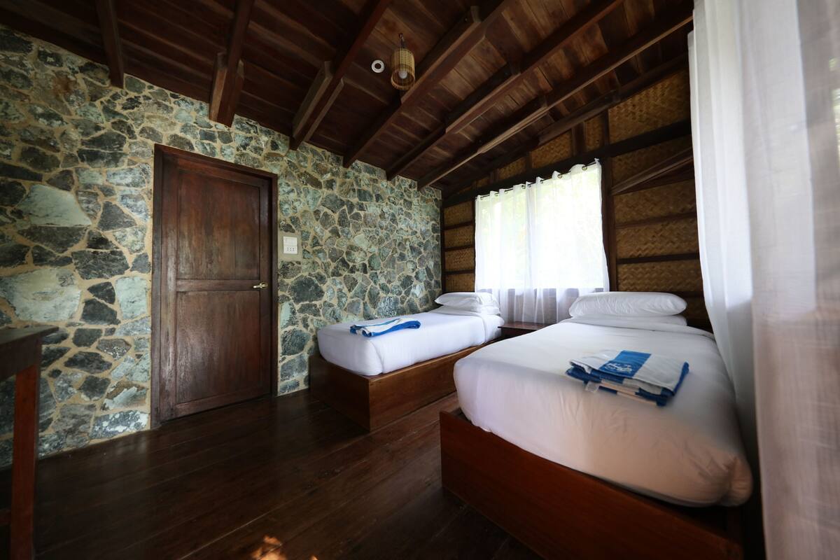 A room with stone walls, wooden floors, two beds, a wooden door, and a window with curtains.