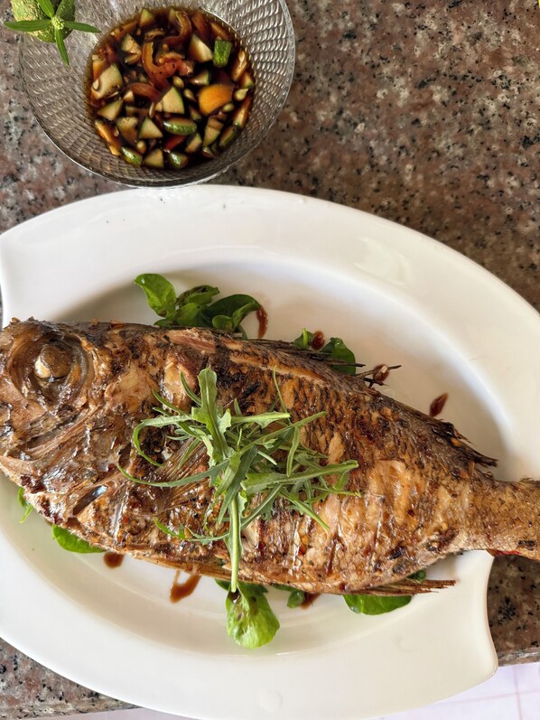 Un pescado entero a la parrilla sobre un plato blanco, acompañado de verduras mixtas y un recipiente pequeño con salsa.