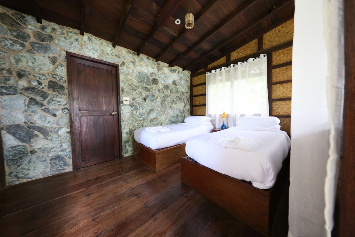 A room with stone walls, wooden floors, two beds, a wooden door, and a window with curtains.