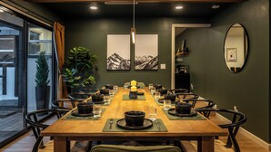 A dining area with a long wooden table set for a meal, surrounded by chairs, with a large potted plant and artwork on the walls. - Summit Haven - Spa - Sauna - Gameroom - 1/4 acre backyard fenced-in (Big Bear Lake)