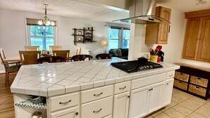 Une cuisine dotée d’un grand îlot central, de meubles blancs et d’une cuisinière à gaz. Deux fenêtres laissent entrer la lumière naturelle, et un coin repas comprend des chaises et une table.