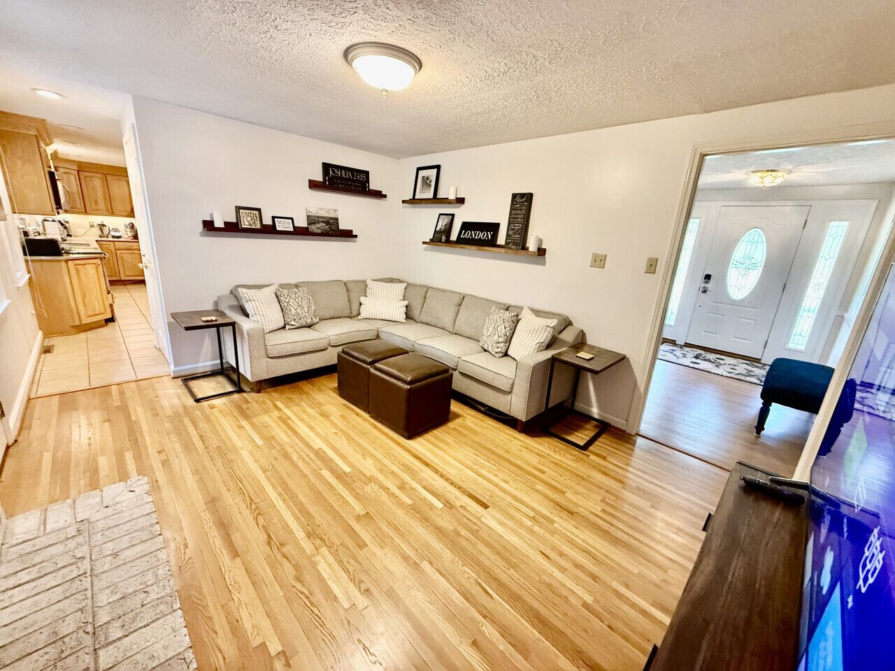 Ein Wohnzimmer mit Ecksofa, Couchtisch und TV-Schrank. Regale mit gerahmten Bildern und eine Tür zum Küchenbereich.