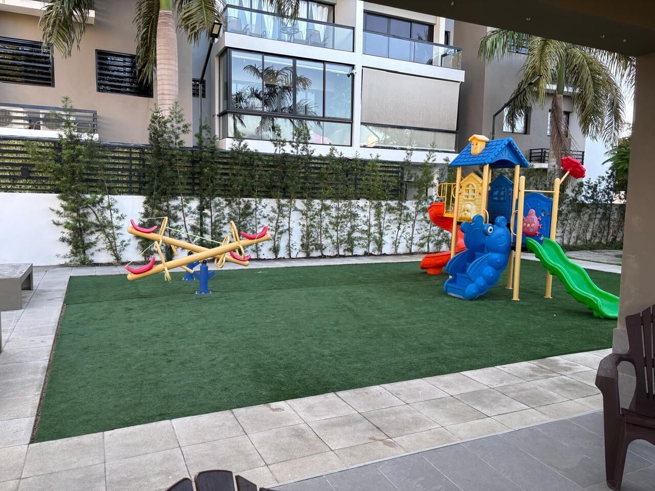 A playground with a seesaw and slides, surrounded by a green artificial turf and a white wall with a black railing.