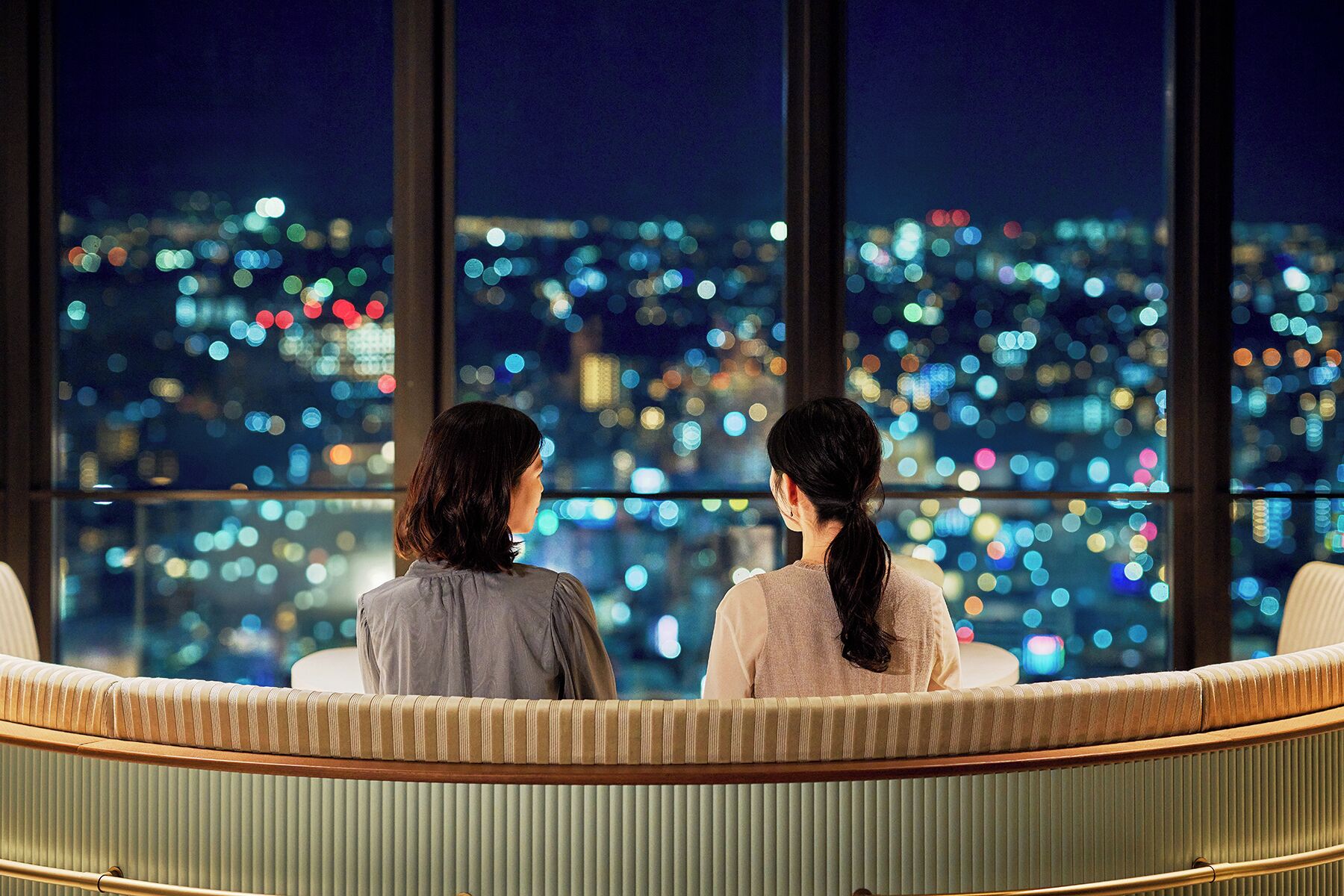 Two people sitting on a curved bench with a cityscape view through large windows.