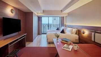A hotel room with a large flat-screen TV, a bed with pillows, a bedside table with a book and a mug, and a view of the city through large windows.