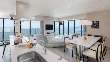 Cozinha moderna com uma ilha grande, área de jantar com mesa redonda e sala de estar com televisão e grandes janelas com vista para a paisagem urbana.