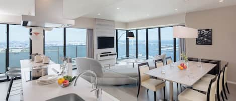 A modern kitchen with a large island, dining area with round table, and a living space with a TV and large windows overlooking a cityscape.