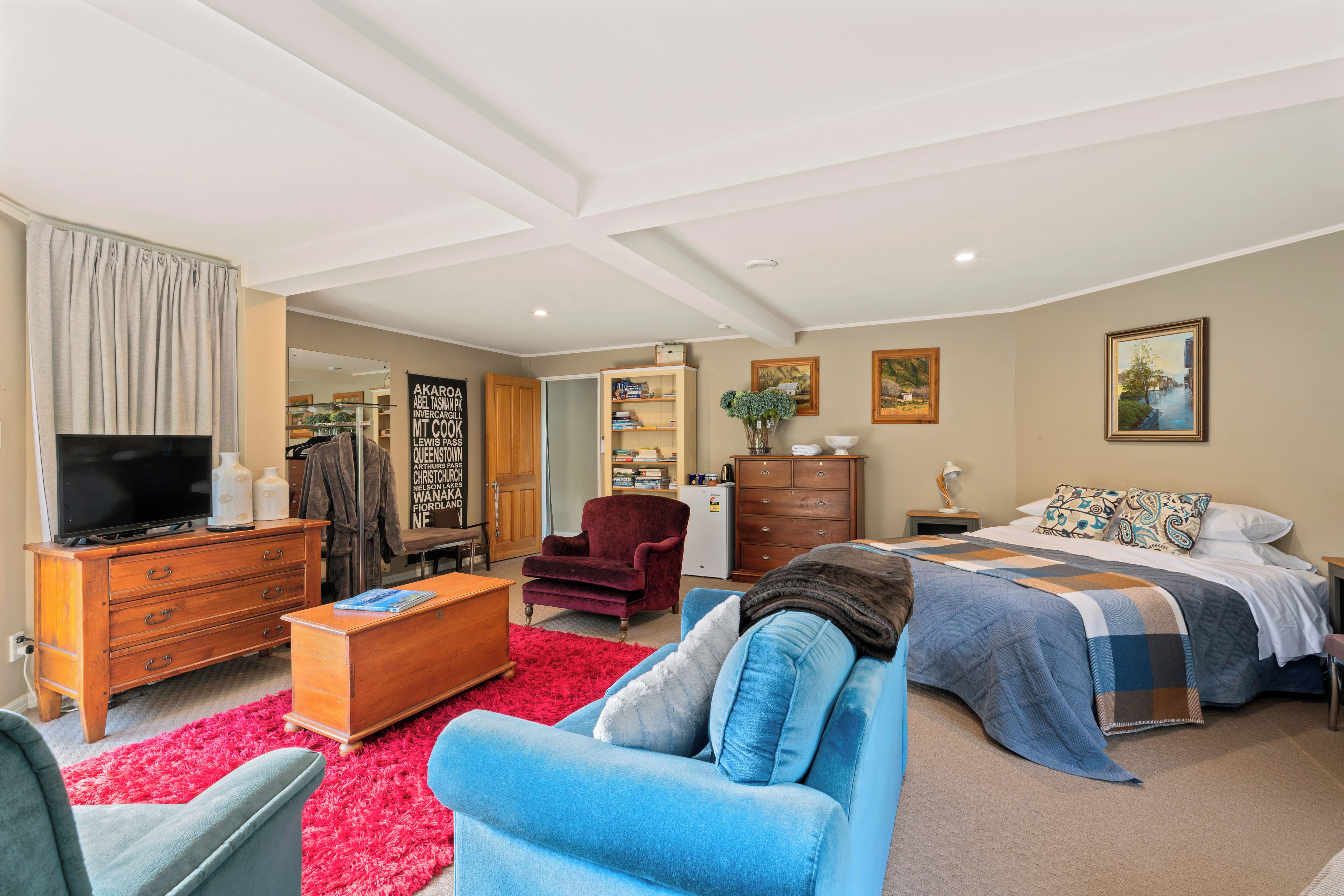 A spacious bedroom with a large bed, a blue sofa, a wooden dresser, a red armchair, a television, and a red shaggy rug.