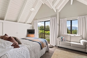 A bedroom with a large bed, a sofa, and a TV. There is a window with curtains and a view of greenery.