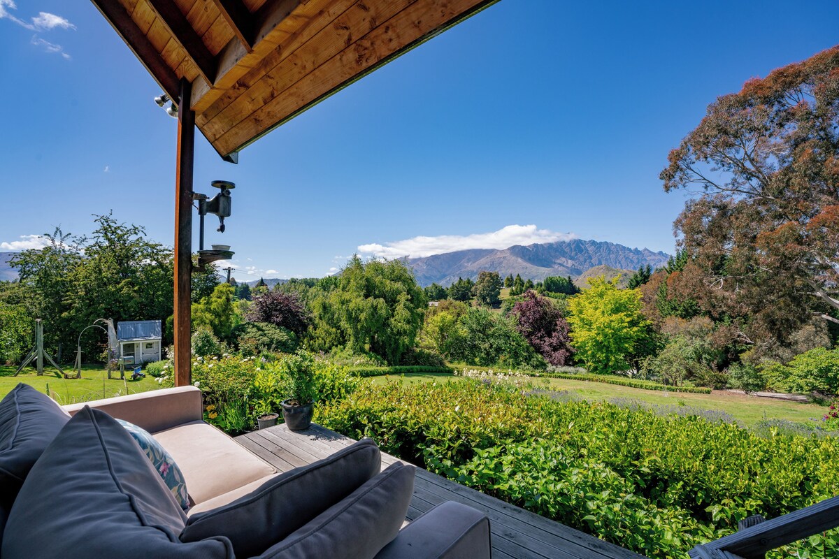 A wooden deck with outdoor seating, overlooking a lush garden and a mountain range.