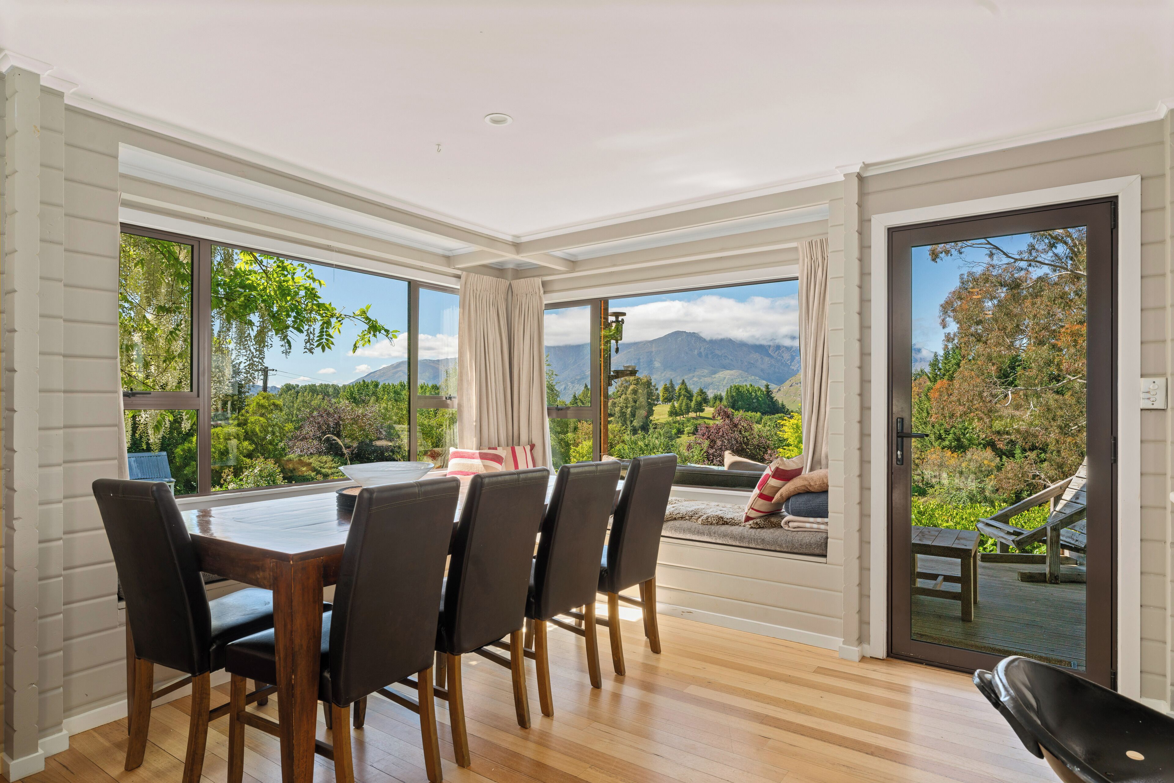 Un espace repas comprenant une table et des chaises, de grandes fenêtres offrant une vue sur les montagnes et la verdure, et une porte-fenêtre coulissante donnant sur un espace extérieur aménagé pour s’asseoir.
