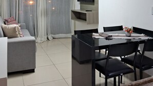 A modern living room with a grey sofa, a dining table with chairs, a flat-screen TV mounted on the wall, and a window with sheer curtains. - Nomar XIX (João Pessoa)