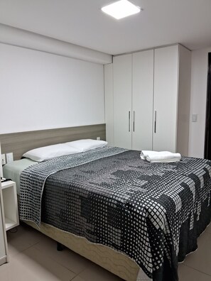 A neatly made bed with a patterned bedspread, a bedside table with a lamp, and a wardrobe with sliding doors. - Nomar XIX (João Pessoa)
