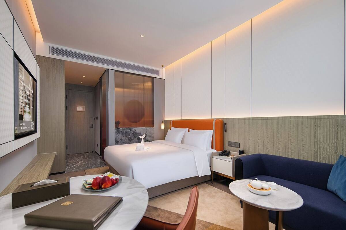 A modern hotel room with a large bed, a sofa, a desk, and a small table. There is a door leading to a bathroom and a hallway.