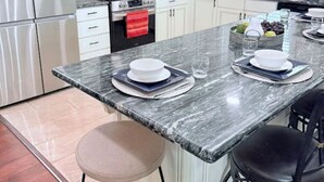 Une cuisine moderne avec des armoires blanches, un réfrigérateur en acier inoxydable, une cuisinière à gaz et un comptoir en marbre. On y trouve deux tabourets de bar et une fenêtre qui laisse entrer la lumière naturelle.