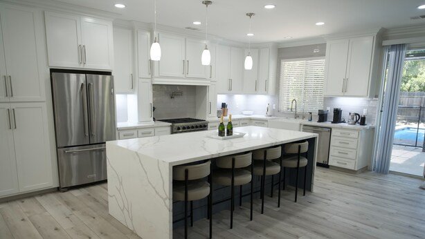 A modern kitchen with white cabinets, a stainless steel refrigerator, a large island with bar stools, and a view of a backyard.