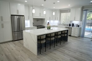Una cocina moderna con armarios blancos, un refrigerador de acero inoxidable, una isla grande con taburetes de bar y vistas a un jardín trasero.