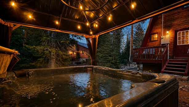 A hot tub area with a wooden deck, a covered seating area, and a wooden cabin in the background.