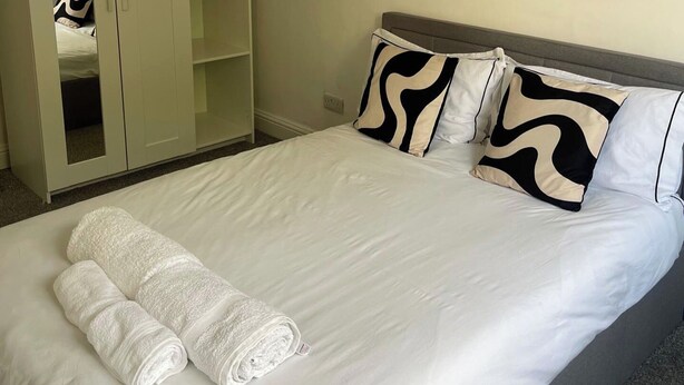 A neatly made bed with white linens and two rolled towels, a grey headboard, and two patterned pillows.