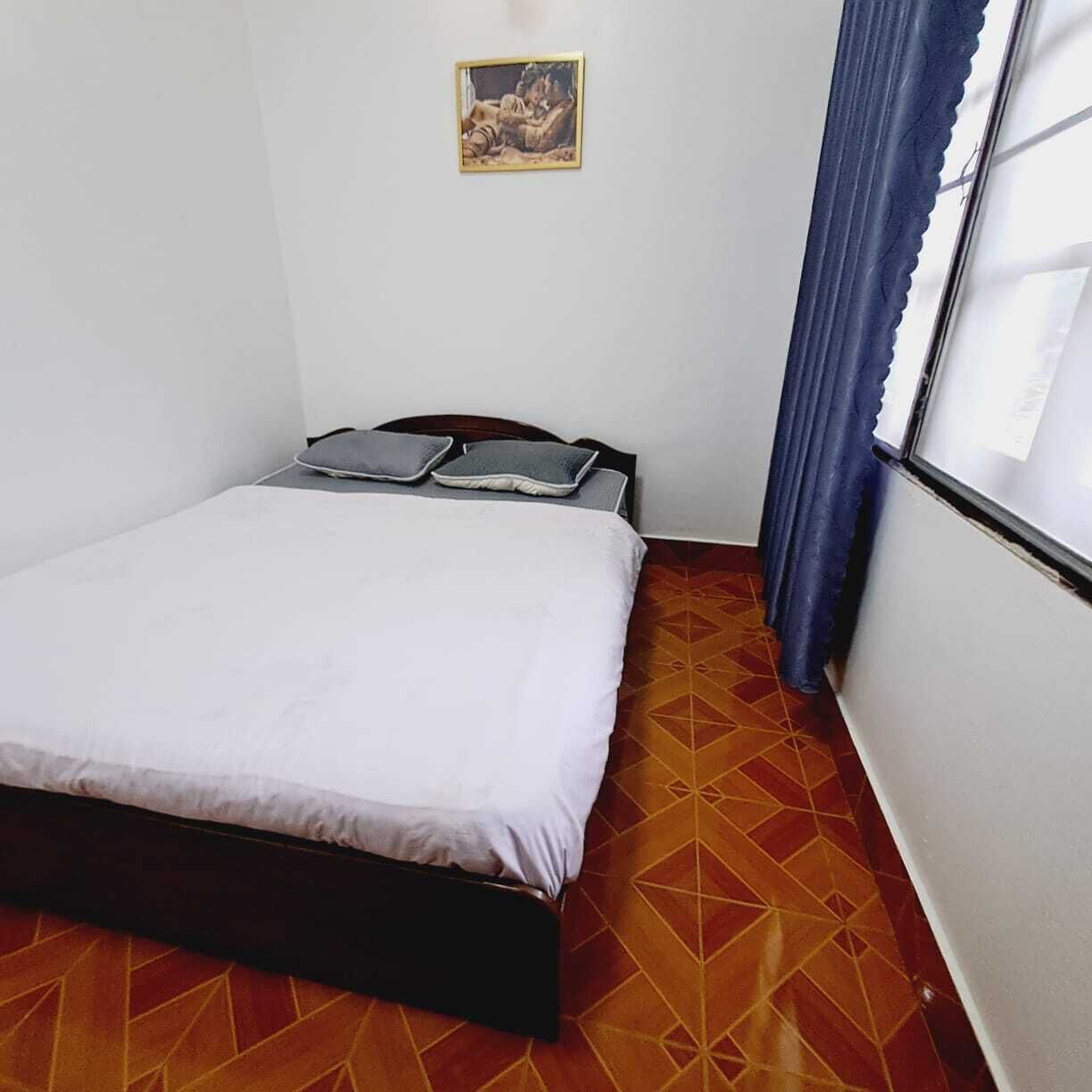 Una cama individual con colchón blanco y estructura de madera oscura, un cuadro pequeño en la pared y una ventana con cortinas azules.