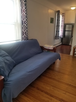 Een woonkamer met een bank, een kleine tafel en een keukenhoek met een koelkast.