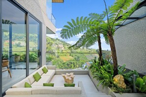 A modern balcony with a glass wall, a white sofa with green cushions, a wooden coffee table, and a fern plant. - Dago Escape Villa by Kozystay - Bandung (Cimenyan)