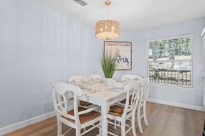 A dining area with a white table set for four, white chairs with woven seats, a hanging wicker chandelier, and a framed picture of a plant. - Polished Condo with Lake Access, Community Outdoor Pool, Mini Golf & Free Attraction Tickets (Kimberling City)