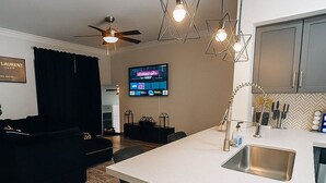 A modern kitchen with a white countertop, stainless steel sink, and a black trash bin. There is a ceiling fan, a black couch, and a television mounted on the wall. - Modern Executive Suite- NRG, Medical Center, Downtown Houston (Houston)