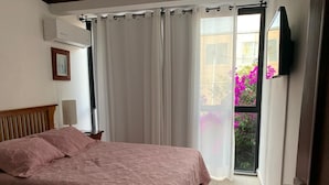Un dormitorio con cama, televisor montado en la pared, ventana con cortinas y balcón con flores.