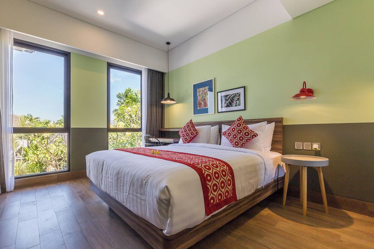 A modern hotel room with a large bed, a small round table, and a view of greenery through the window.