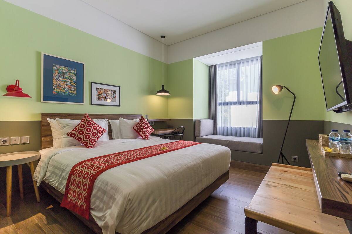 A hotel room with a bed, bedside table, window, artwork, and a desk with a lamp.