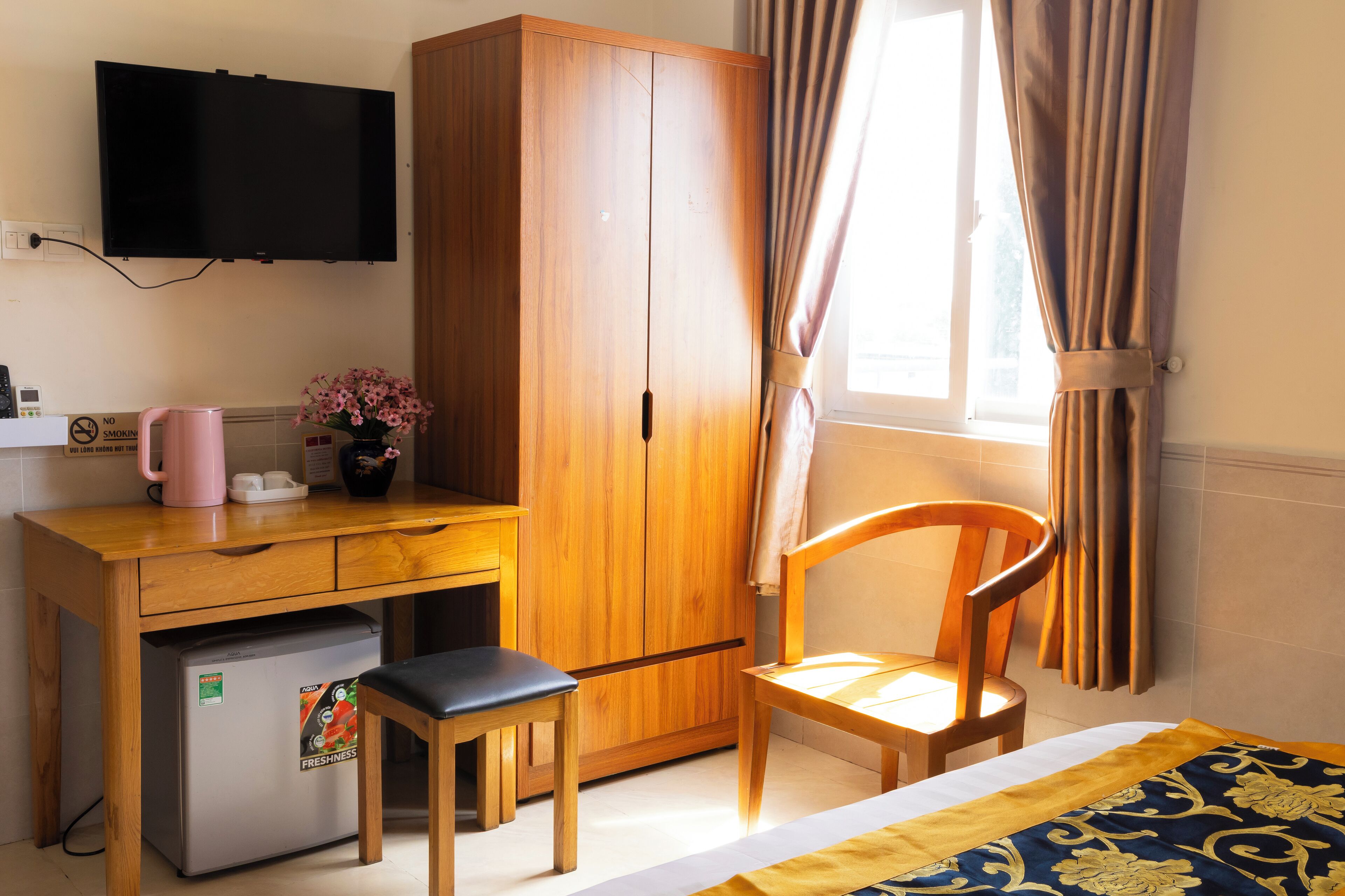 A hotel room with a wooden desk, a chair, a bed with a patterned bedspread, a television mounted on the wall, and a window with curtains.