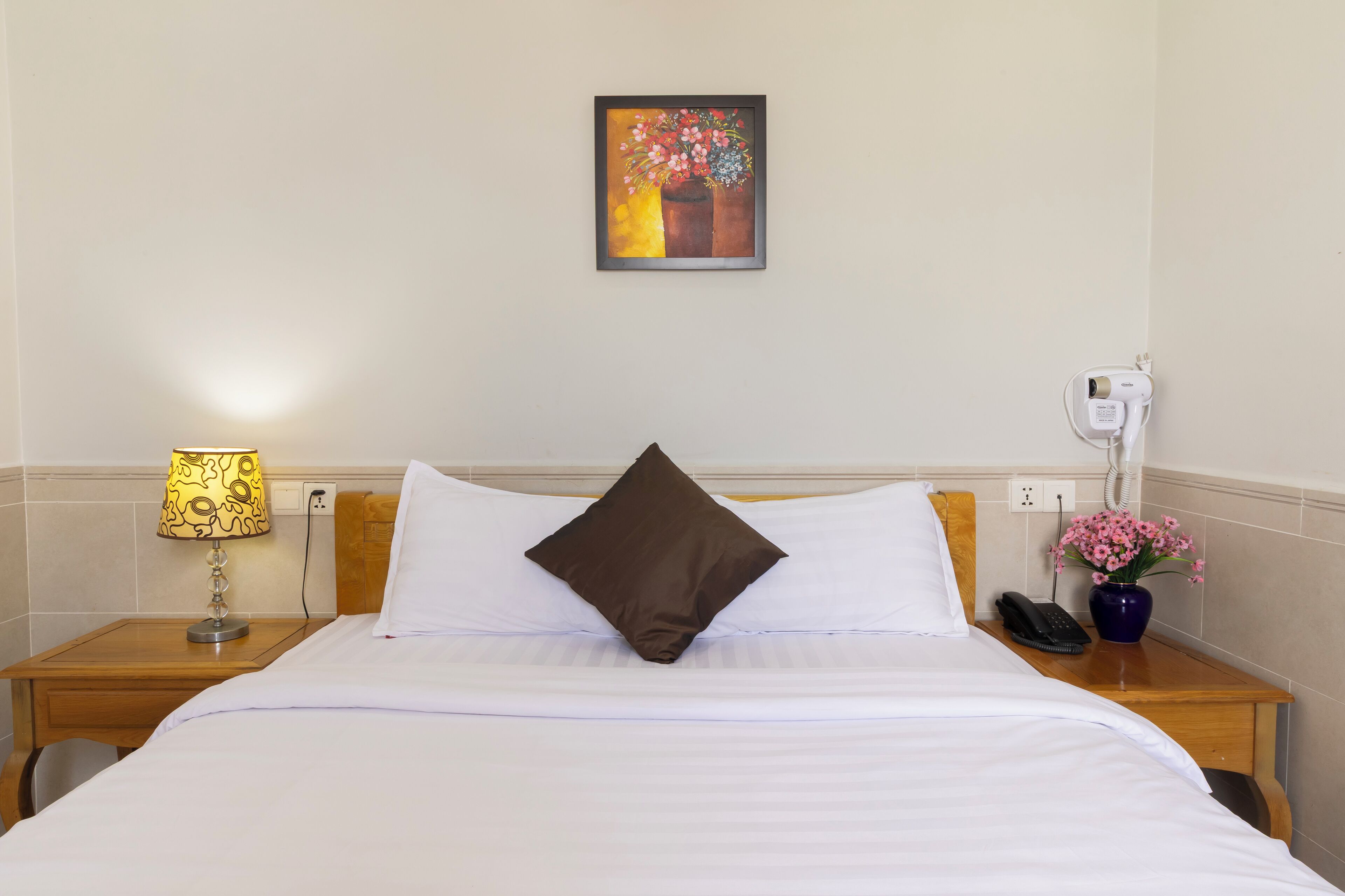 A hotel room with a bed, a nightstand with a lamp, a phone, and a vase with flowers.