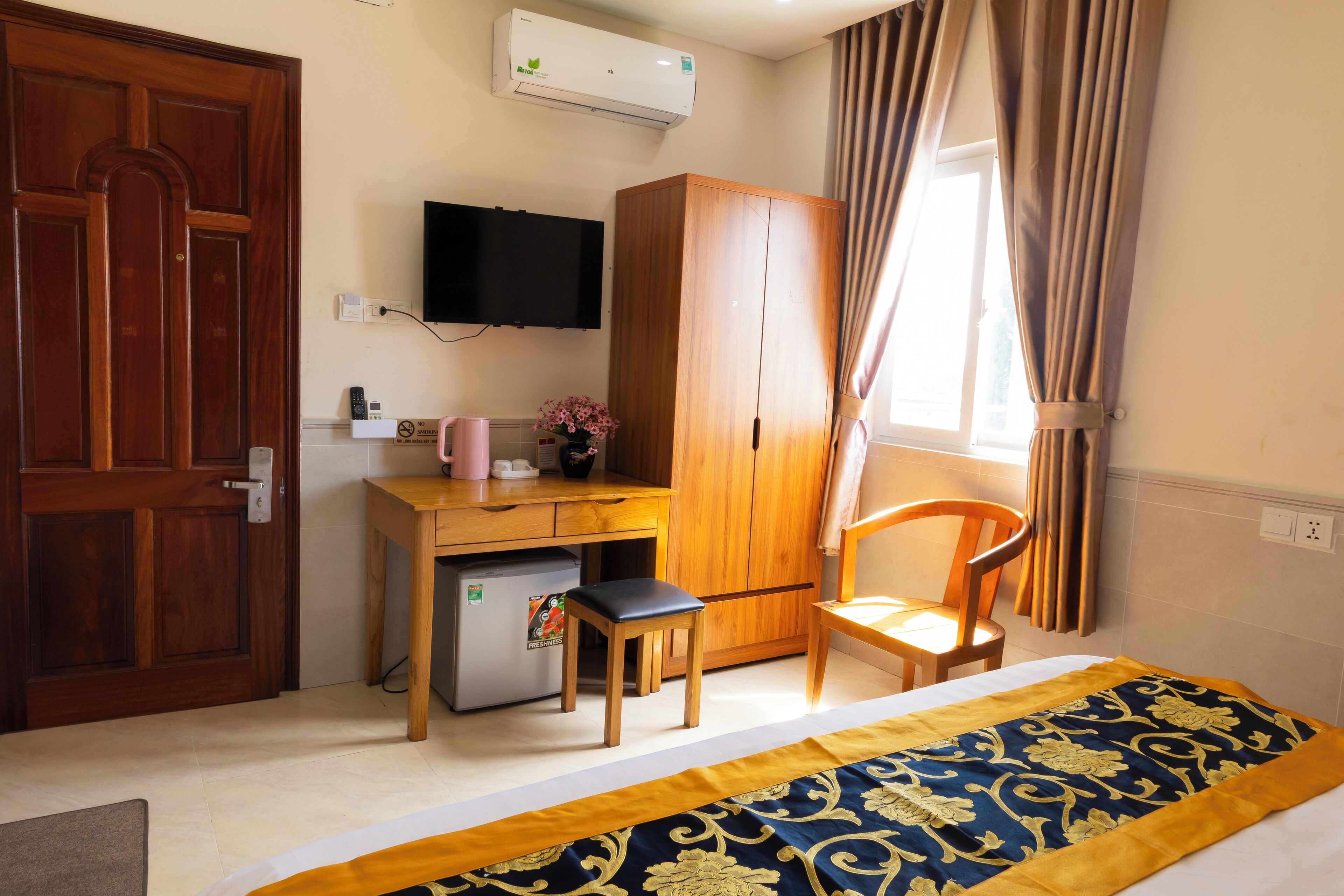 A hotel room with a wooden desk, a chair, a bed with a patterned bedspread, a wardrobe, a television mounted on the wall, and a window with curtains.