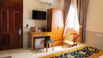 A hotel room with a wooden desk, a chair, a bed with a patterned bedspread, a wardrobe, a television mounted on the wall, and a window with curtains.
