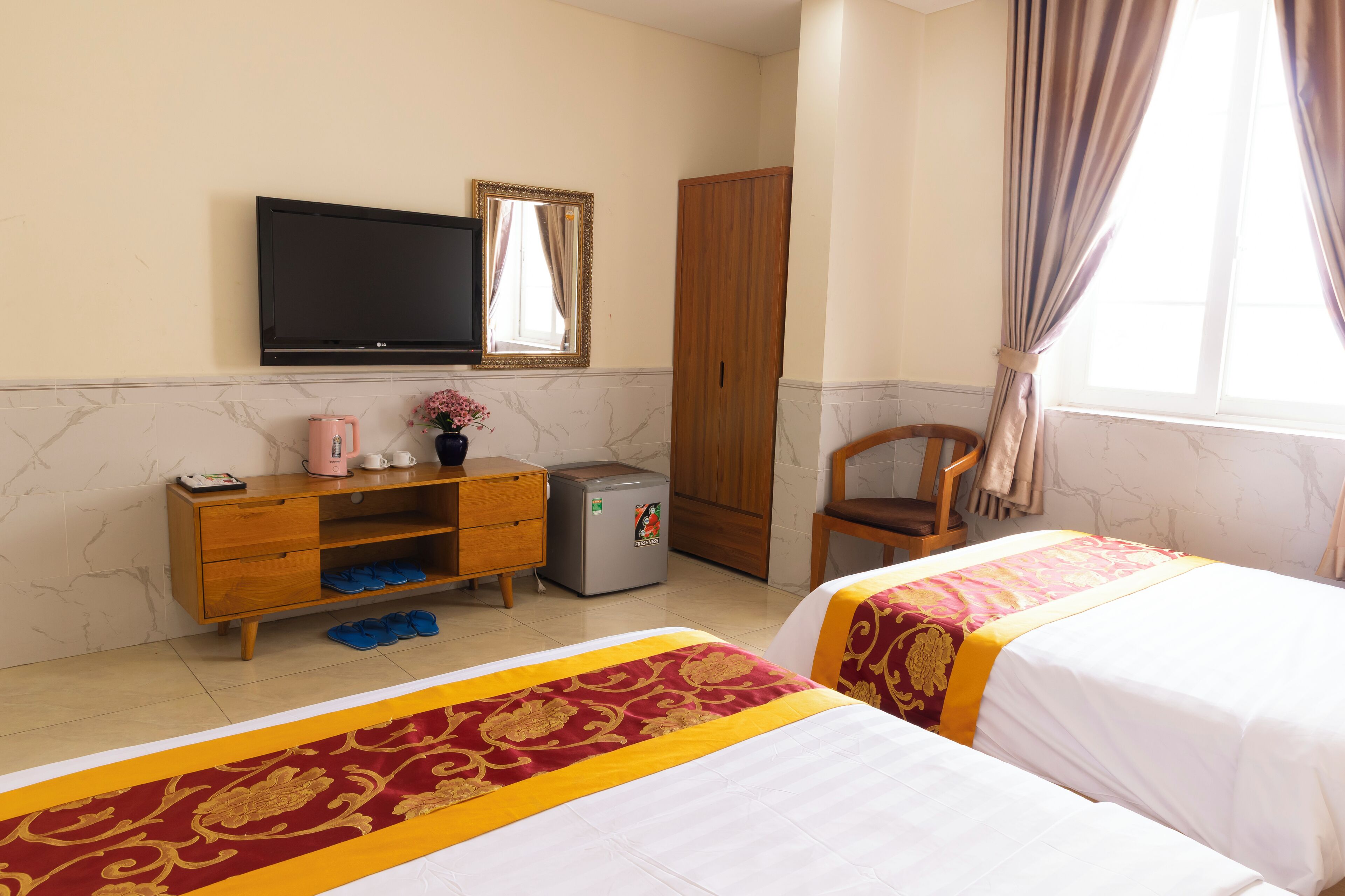 A hotel room with two beds, a TV, a wardrobe, a small refrigerator, and a wooden chair.