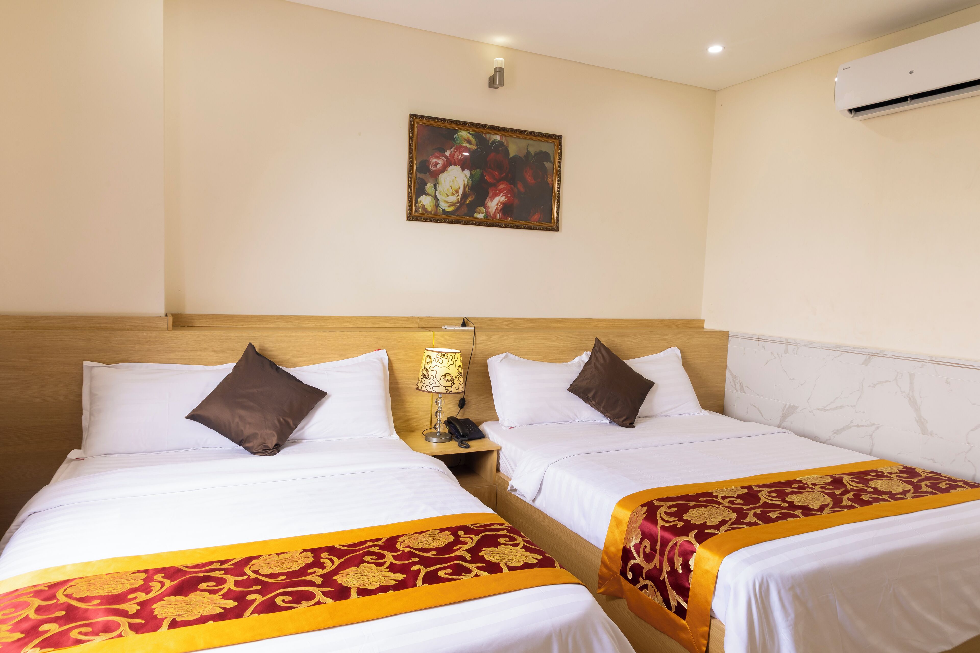 Une chambre d’hôtel avec deux lits, chacun agrémenté de draps blancs et d’une couvre-lit à motifs rouges et dorés. Un tableau est accroché au mur au-dessus des lits, et un appareil de climatisation est fixé au mur, à droite.