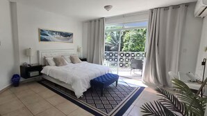 Un dormitorio con cama, mesita de noche, lámpara, un banco azul, alfombra estampada y balcón con cortinas.