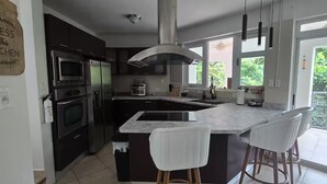 Una cocina moderna con gabinetes oscuros, electrodomésticos de acero inoxidable, una isla grande con taburetes de bar y vistas a zonas verdes a través de amplios ventanales.
