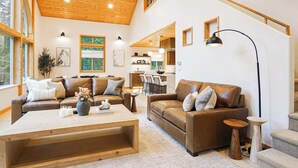 A spacious living area with a high ceiling, wooden beams, and a staircase leading to an upper level. There are two leather sofas, a coffee table, and a dining area with chairs.