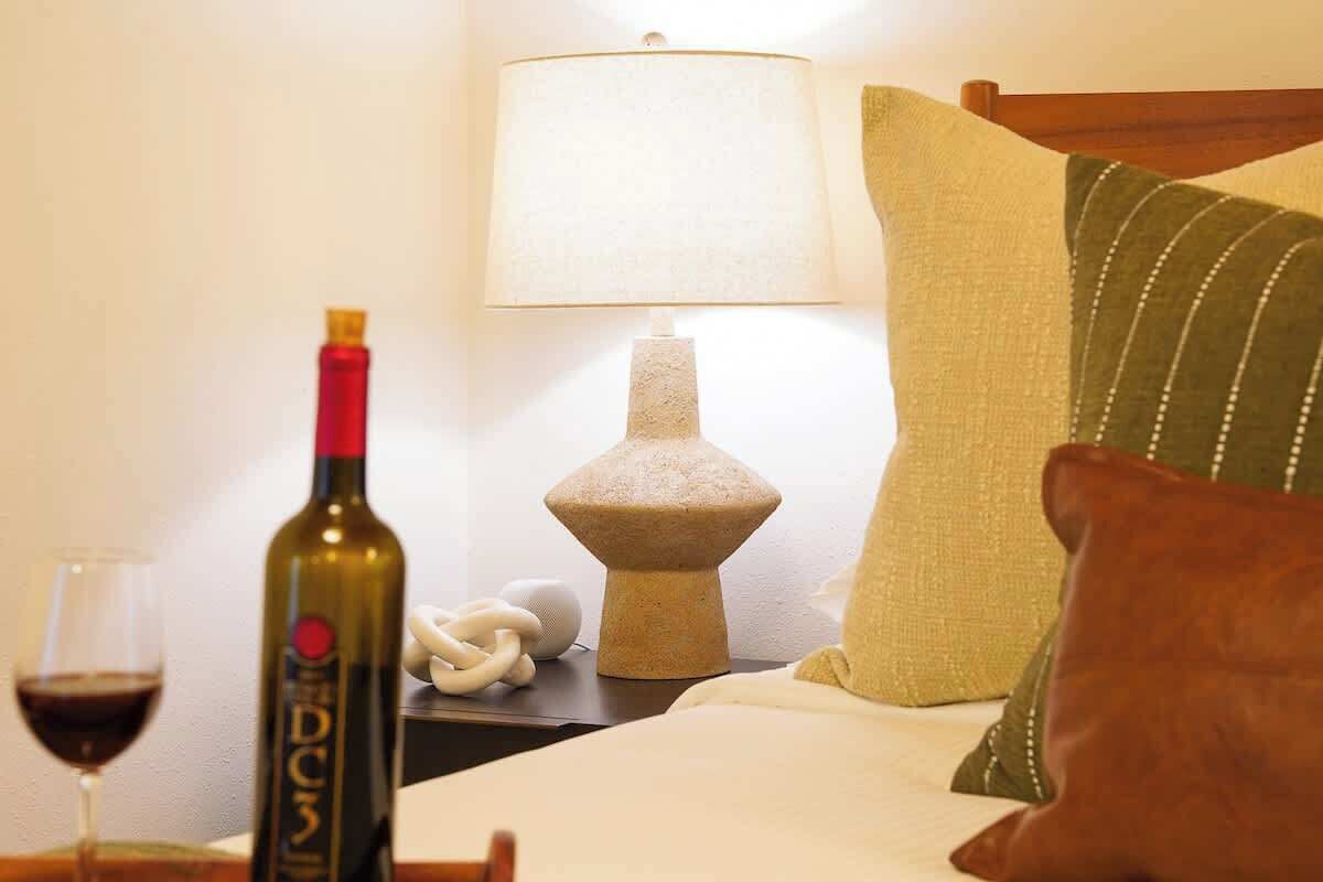 A hotel room with a bed, a lamp, a bottle of wine, and a glass of wine on the nightstand.